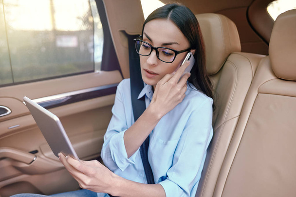 serious businesswoman talking on smartphone and holding digital tablet in the car - Фото, изображение