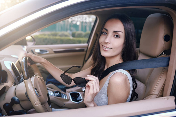 portrait of brunette woman holding sunglasses and looking at the camera while sitting in the car - Foto, Bild