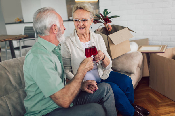 Wesoły starszy para Przeprowadzka do nowego domu uśmiechając się wzajemnie i pić wino. - Zdjęcie, obraz
