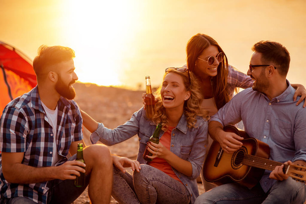 Giovani amici felici che fanno picnic sulla spiaggia.Sono tutti felici, si divertono, sorridono e suonano la chitarra
. - Foto, immagini