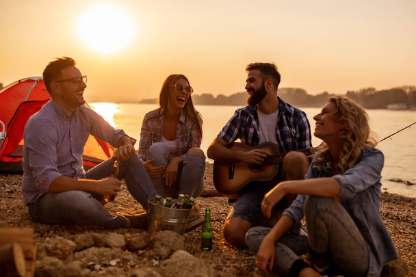 幸せな若い友人がビーチでピクニックを持ちます。彼らがすべて幸せ、楽しんで、笑顔でギターを弾く. - 写真・画像