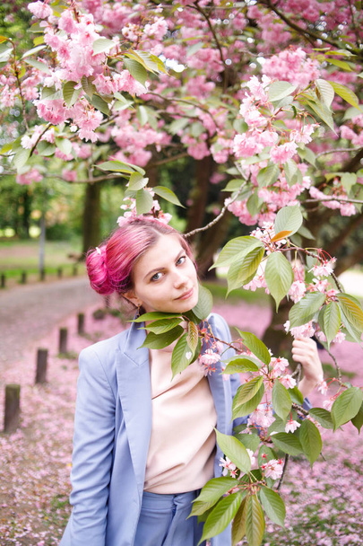 Piękna dziewczyna z różowe włosy w modne niebieski pantsuit stojącą obok kolorowych kwiatów. . Dość miękki model wygląda do aparatu. - Zdjęcie, obraz