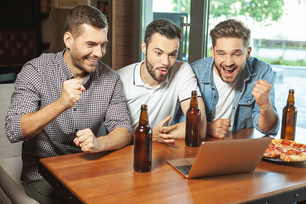 three fans drinking beer and watching sports game on laptop computer in the cafe - Foto, imagen