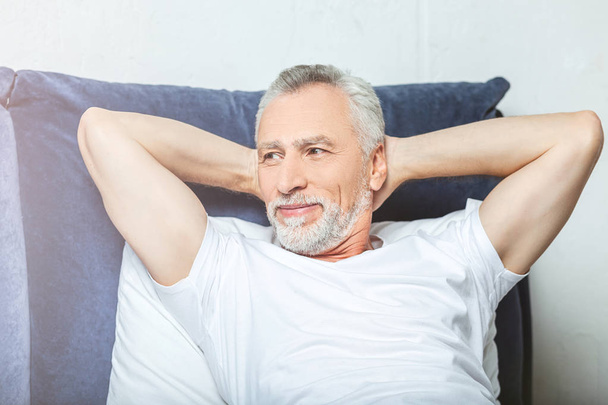 smiling senior man lying in bed, holding hands behind the head and looking away - Photo, Image