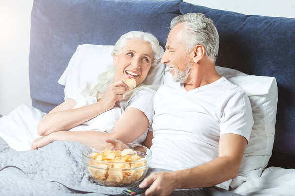 hymyilevä vaimo ja aviomies katsomassa televisiota ja syöminen pelimerkkejä sängyssä
 - Valokuva, kuva