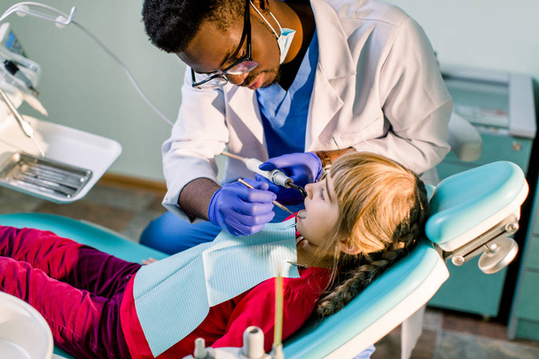 Young beautiful girl in dental office. Children's African-American dentist examines teeth to a child. - Фото, зображення