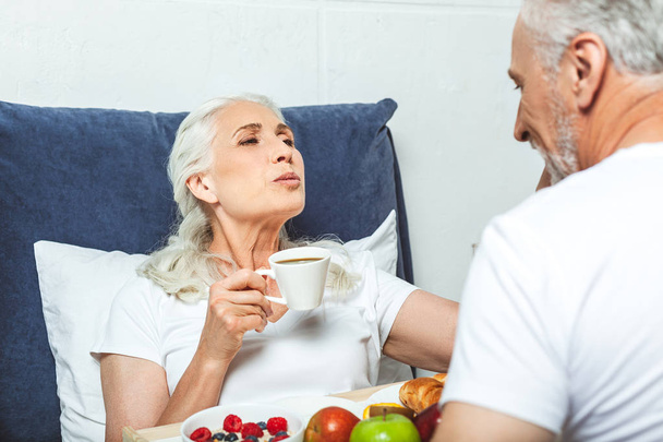 senior woman having breakfast in the bed, holding coffee cup and trying to kiss her husband - Photo, image