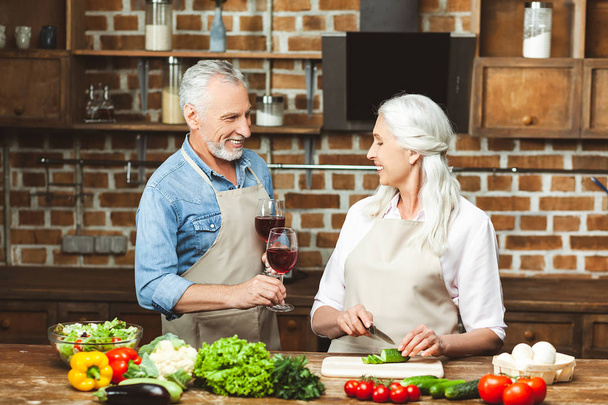 husband holding glasses with red wine while wife preparing salad at the kitchen - Photo, Image