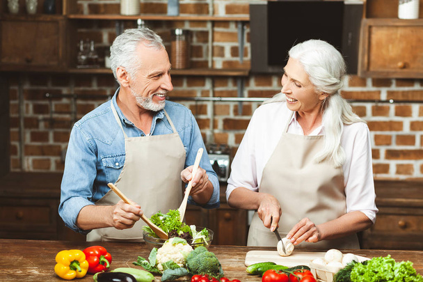 senior couple preparing salad for dinner and looking at each other at the kitchen - Photo, Image
