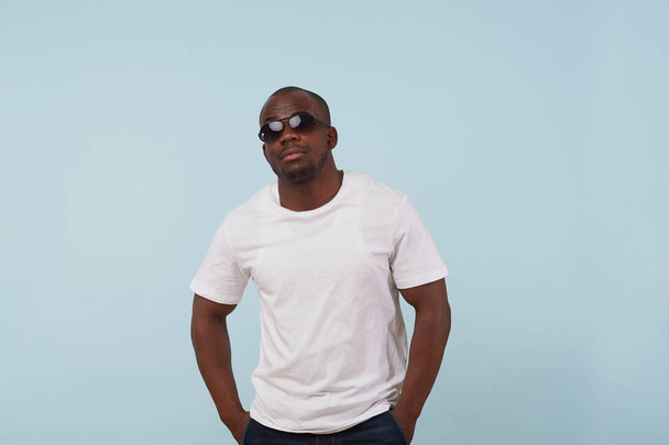 Cool fit black man in white T-shirt and sunglasses is looking at camera against pale blue background. Serious, bold, stubble beard. Hands in pockets. - Fotografie, Obrázek