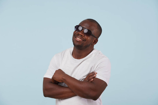 Smiling black man in white T-shirt and sunglasses is looking at camera against pale blue background. Bold, stubble beard. Hands crossed. - Foto, Imagem