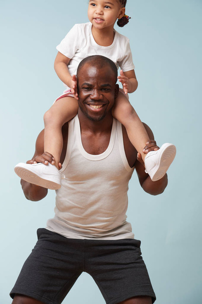 Handsome black young father is doing squats with his cute little daughter riding him. They have fun, laugh together. - Foto, Imagem