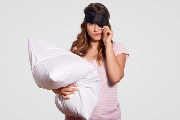 Studio shot of pleasant looking girl covers eyes with eye mask, holds soft pillow, prepares for having sleep in bedroom, dressed in pyjamas, isolated over white background. Relaxation concept - Foto, Bild