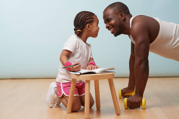 Cute little daughter fitness training her handsome black young father. She is making notes in her paper notebook. Child role play. They are looking into each other's eyes, giggling, having fun. - Foto, Imagen