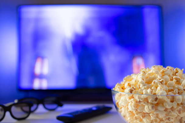 Skleněná mísa popcornu, 3d brýle a dálkové ovládání na pozadí Tv funguje. Večer příjemné sledování filmu nebo televizní seriál doma. - Fotografie, Obrázek