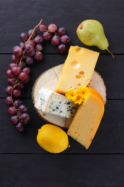 Different types of cheese and grapes on a wooden stand. Slices of cheese and a bunch of grapes on a dark background. Camembert, blue and hard yellow cheese - Foto, immagini