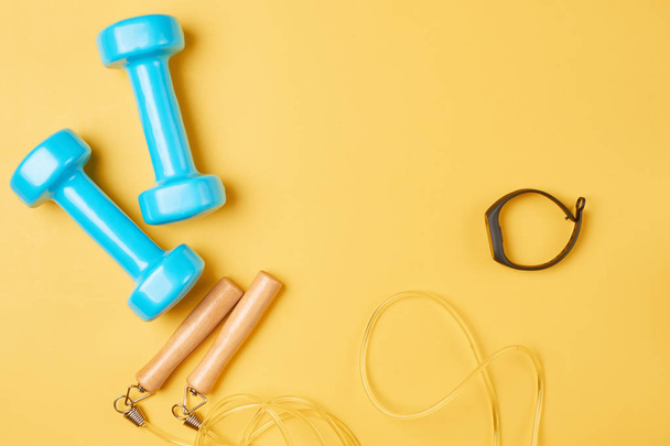 Flat lay composition of blue dumbbells, jump rope and fitness tracker on a yellow background with copy space - Photo, Image