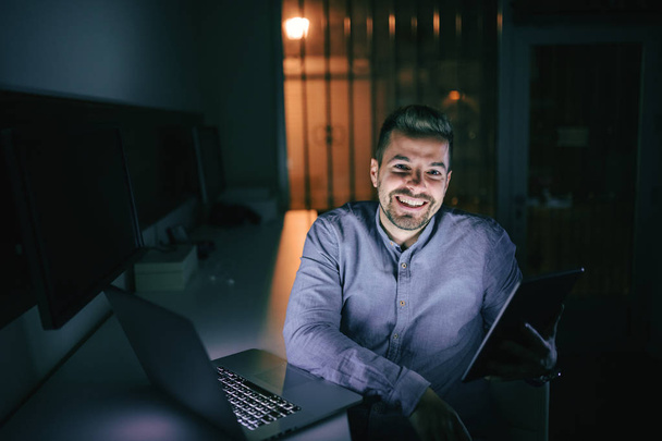 Employee with toothy smile on his face using tablet while sitting in the office late at night. On desk laptop. - Zdjęcie, obraz