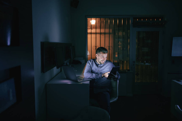 Emplear sonriendo y usando la tableta mientras está sentado en la oficina tarde en la noche. En la computadora portátil de escritorio
. - Foto, imagen