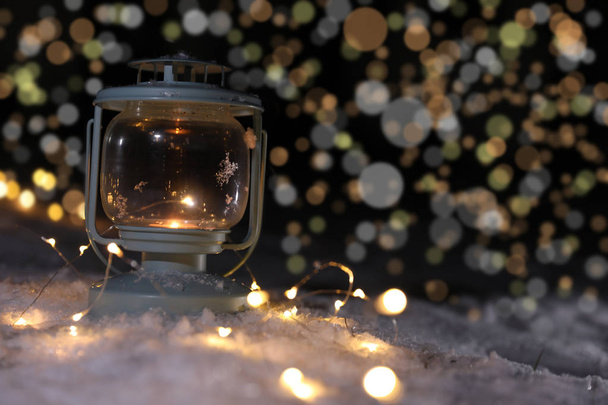 Lantern with burning candle and Christmas lights on snow against blurred background, space for text. Winter decor - Foto, Imagem