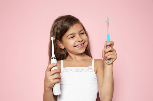 Little girl choosing between manual and electric toothbrushes on color background - Fotó, kép