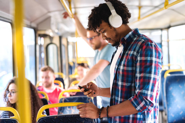 Молодой улыбающийся афроамериканец, слушающий музыку и пользующийся смартфоном во время езды в городском автобусе
. - Фото, изображение