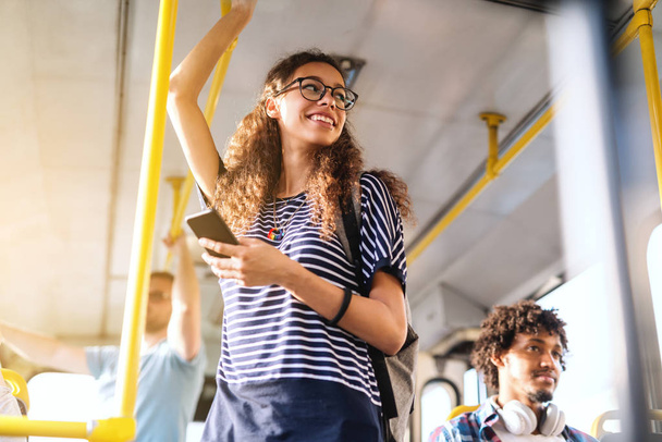 Dziewczyna piękna mieszanej rasy z długie kręcone włosy za pomocą inteligentnego telefonu do czytania lub pisania wiadomości stojąc w autobus miejski. - Zdjęcie, obraz