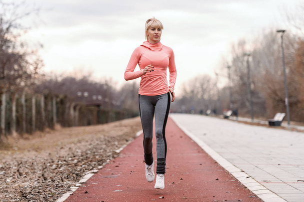 Gorgeous dedicated blonde Caucasian woman in sportswear and with ponytail running on the racetrack. - Photo, Image