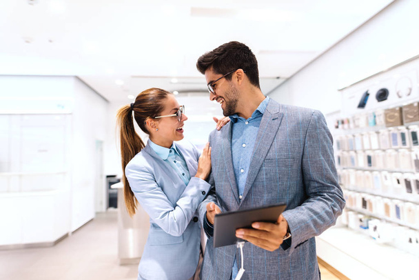 Happy multicultural couple in formal wear buying new tablet while standing in tech store. Man holding tablet while woman leaning on him. - Фото, изображение