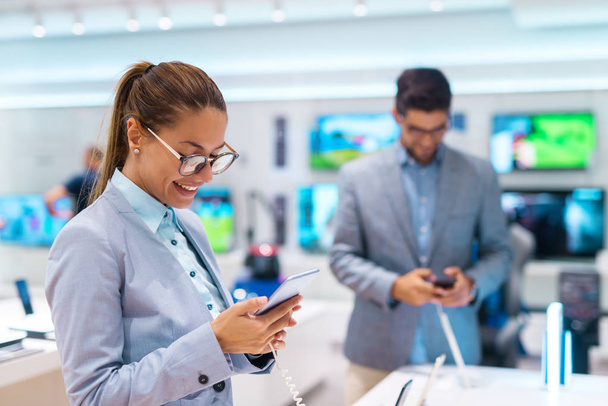 Happy beautiful Caucasian woman with ponytail and dressed in suit buying new smart phone. Tech store interior. - Φωτογραφία, εικόνα