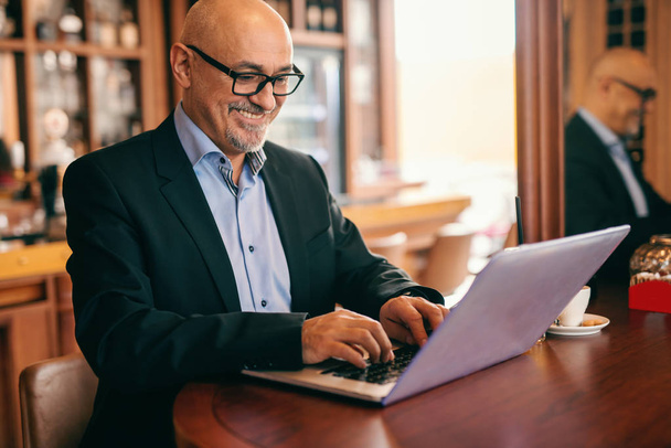 Старший бородатый бизнесмен в костюме использует ноутбук для работы, сидя в кафетерии. На столе кофе
. - Фото, изображение