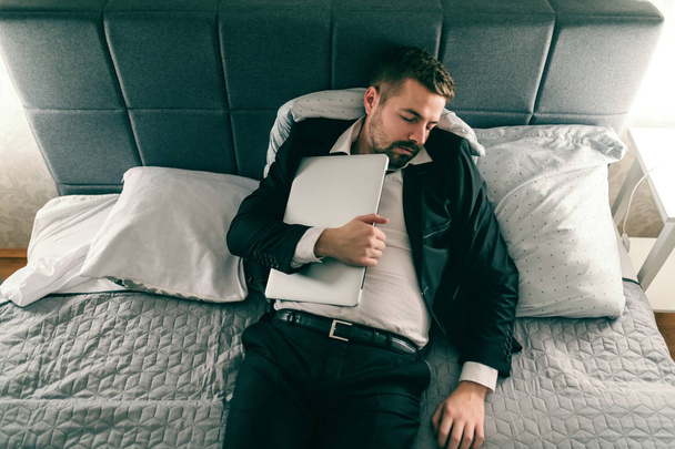Túlhajszolt üzletember ruha alszik az ágyon és laptop tartja a mellét. - Fotó, kép