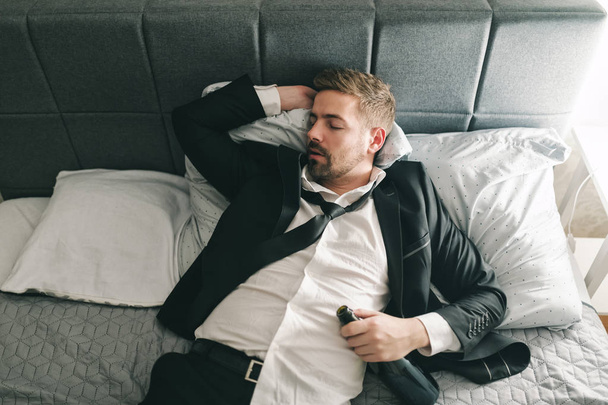 Вид зверху п'яного бізнесмена в костюмі лежить у ліжку і спить з пляшкою алкоголю в руці. Концепція зловживання алкоголем
. - Фото, зображення