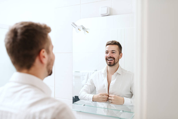 Lachende knappe zakenman verkleden voor werk terwijl je in de badkamer voor spiegel. - Foto, afbeelding
