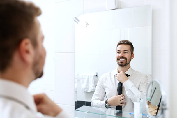 Empresário sorrindo colocando gravata ao olhar no espelho e de pé no banheiro
. - Foto, Imagem