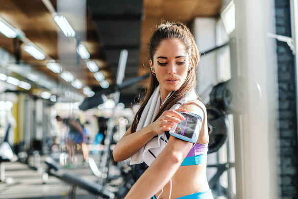 Serious sporty woman in sportswear and towel around neck putting smart phone in phone case on hear arm. In background exercise equipment. Gym interior. - 写真・画像