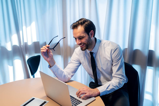 Χαμογελαστός επιτυχημένος εταιρικός επιχειρηματίας χρησιμοποιώντας φορητό υπολογιστή ενώ καθόταν στο γραφείο του. Στο χέρι γυαλιά. - Φωτογραφία, εικόνα