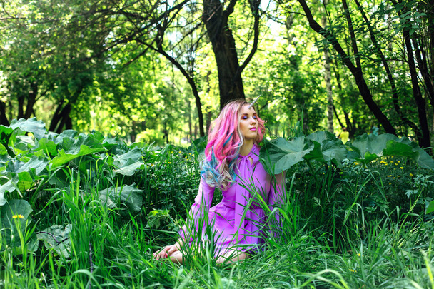 Fairy girl unicorn with rainbow hair and shiny makeup sitting on the grass in the forest. - Valokuva, kuva
