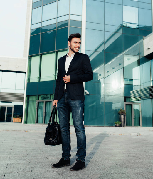 handsome young businessman with a beard and in a business suit standing on the street against the background of the office building next to a comfortable stylish leather bag. - Fotografie, Obrázek