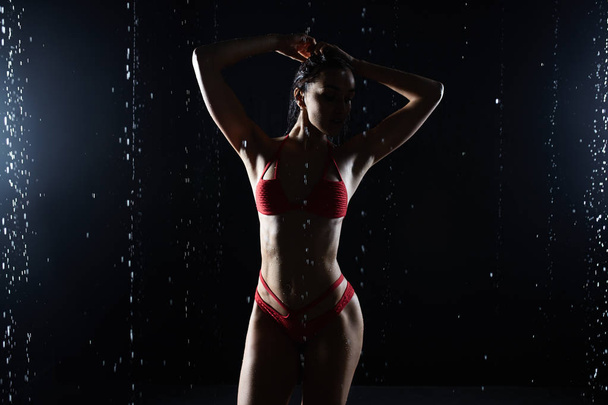 Beautiful young girl in red sportswear poses in aqua studio. Drops of water spread about her fitness body. The perfect figure on the background of water splashes - Photo, image