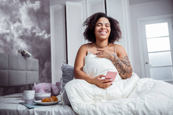 Laughing curly woman wearing pajamas while sitting in bed - Photo, image