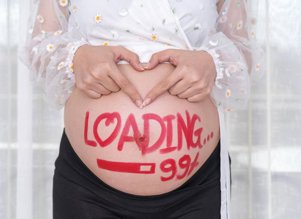 těhotná žena drží za ruce ve tvaru srdce s malovanými štětec slovem - načítání 99 % na břiše - Fotografie, Obrázek