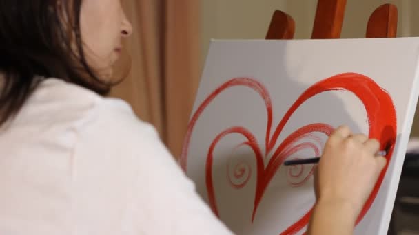 young woman artist painting a red heart with acrylic paint on a white canvas on the easel in her art studio - Footage, Video