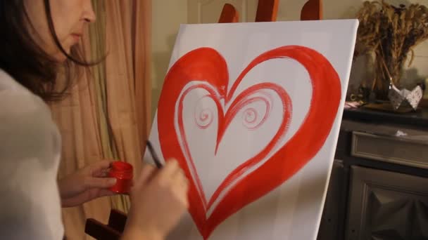 jonge vrouw kunstenaar een rood hart schilderen met acrylverf op een wit doek op de ezel in haar kunststudio - Video
