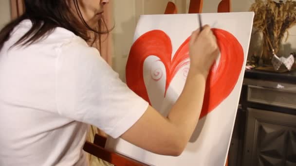 νεαρή γυναίκα καλλιτέχνης ζωγραφίζει μια κόκκινη καρδιά με ακρυλικό χρώμα σε ένα λευκό καμβά σε καβαλέτο στο ατελιέ της τέχνης - Πλάνα, βίντεο