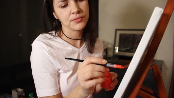 νεαρή γυναίκα καλλιτέχνης ζωγραφίζει μια κόκκινη καρδιά με ακρυλικό χρώμα σε ένα λευκό καμβά σε καβαλέτο στο ατελιέ της τέχνης - Πλάνα, βίντεο