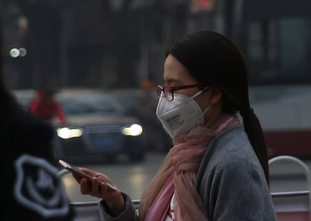A pedestrian wearing a face mask against air pollution uses her smartphone on a street in heavy smog in Beijing, China, 16 March 2016 - Foto, afbeelding