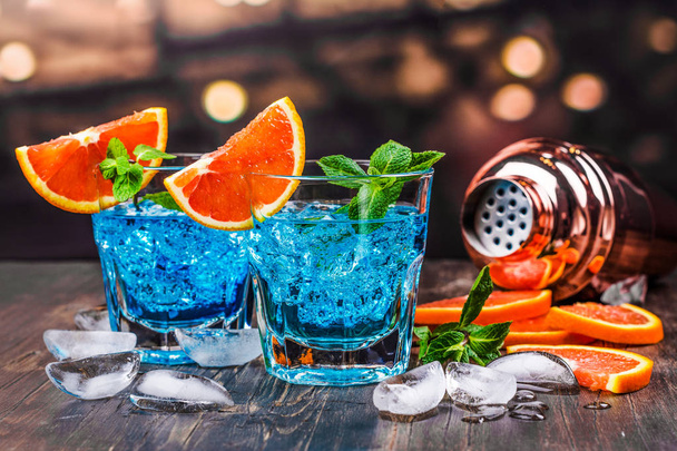 Blue lagoon cocktail in a glass on blured bar background. Blue hawaii cocktail with ice cubes and orange slices - 写真・画像