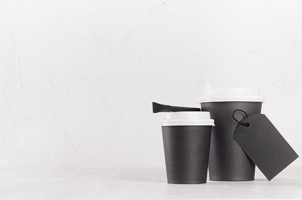 Coffee mockup - two black paper cups with white caps, blank label and sugar bag on white wood table with copy space, coffee shop interior. Modern elegant concept for branding identity, advertising, design. - Foto, Imagem