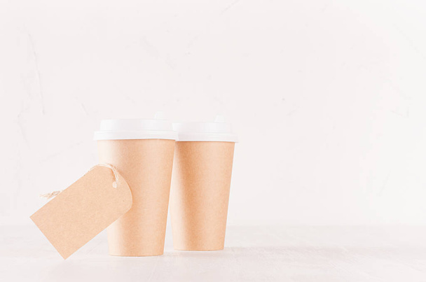 Coffee packing mockup - two craft brown paper cups with white caps and blank label on white wood board with copy space, coffee shop interior. Modern elegant concept for branding identity, advertising, design. - Foto, Imagem